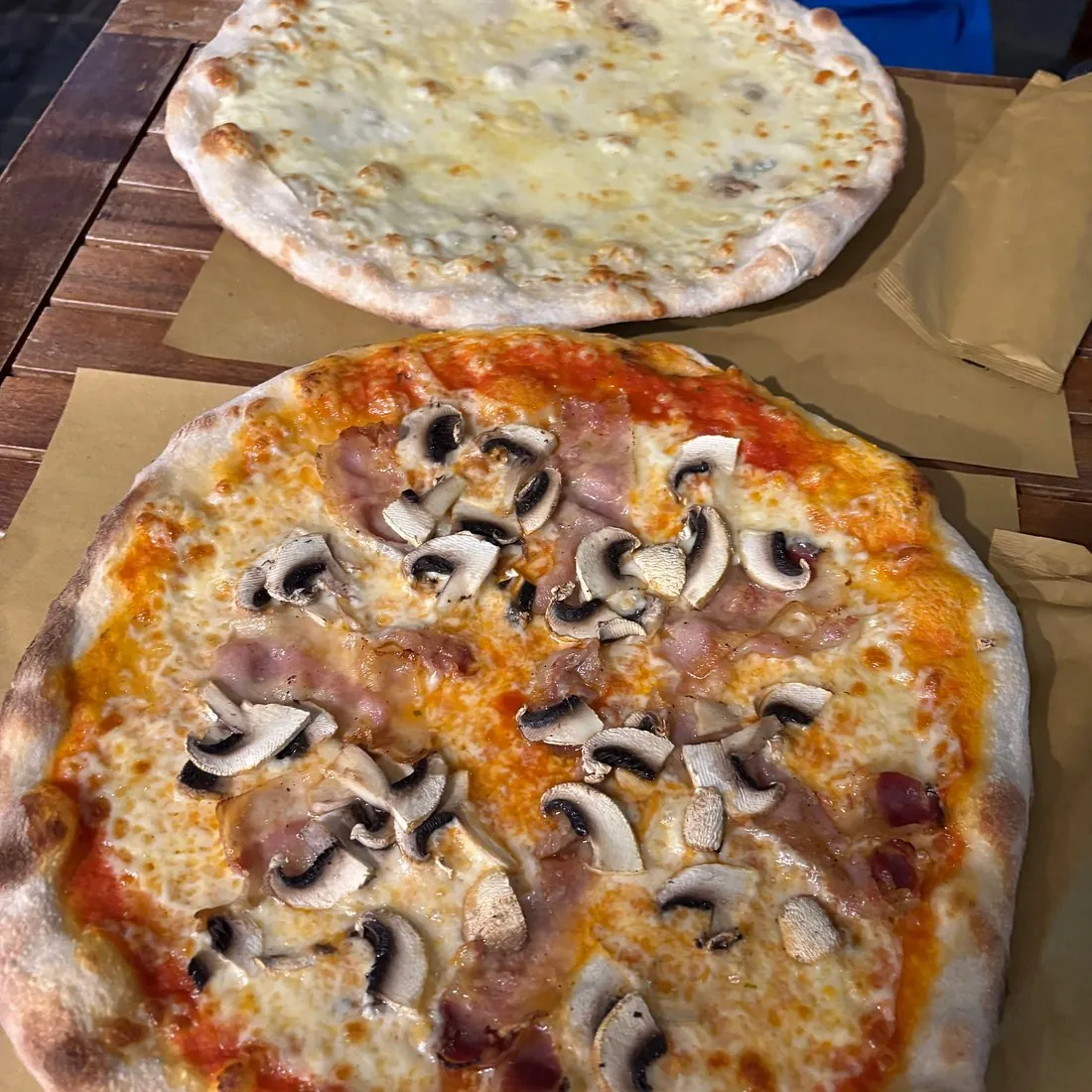 Due pinse appena uscite dal forno su un tavolo di legno, una bianca ai formaggi e una con funghi champignon