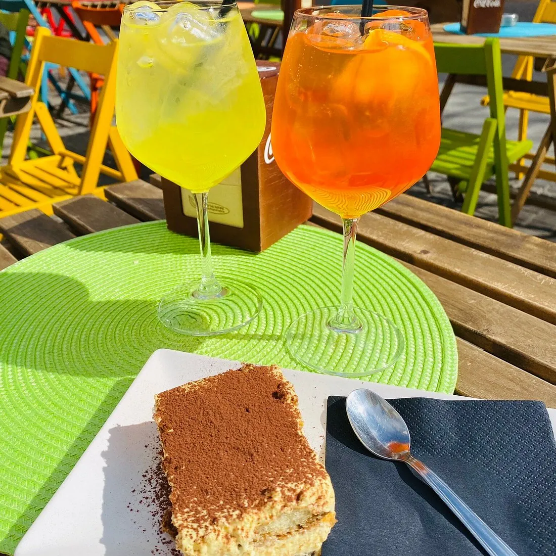 Due spritz, uno giallo e uno arancione, accanto a una fetta di tiramisù su un tavolino esterno